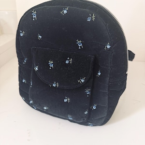 May include: A black velvet backpack featuring a small blue floral pattern, a front pocket with a flap closure, and a silver-toned metal zipper that runs along the top and sides of the bag. The fabric has a quilted texture throughout the exterior.