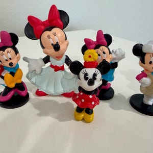 May include: A collection of Minnie Mouse figurines in various poses. The figurines feature Minnie in different outfits, including a red polka dot dress, a cowgirl outfit, and a dress with a red bow. The figurines are on a white surface.