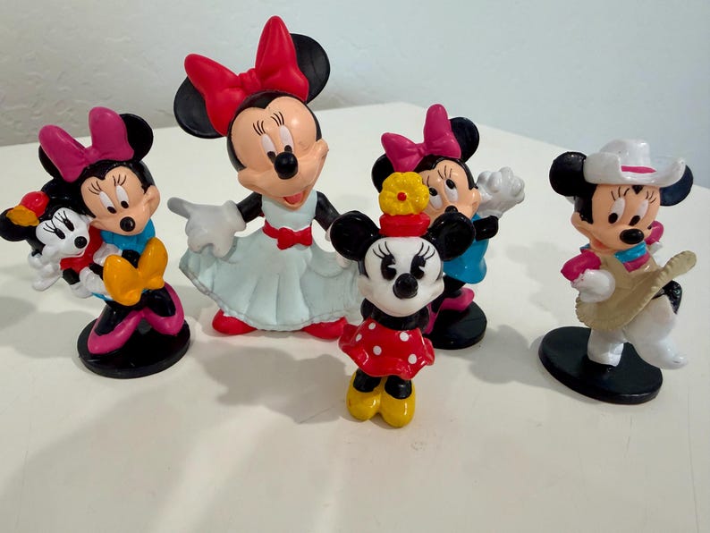 May include: A collection of Minnie Mouse figurines in various poses. The figurines feature Minnie in different outfits, including a red polka dot dress, a cowgirl outfit, and a dress with a red bow. The figurines are on a white surface.