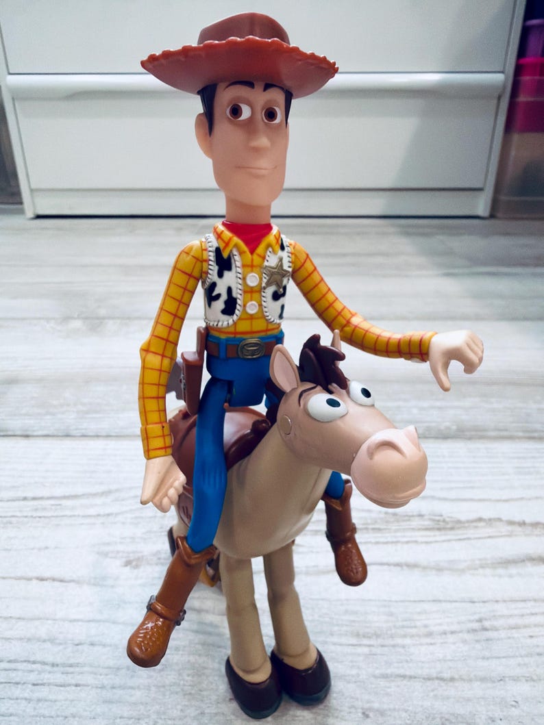 May include: A Woody action figure from the Toy Story franchise, wearing a cowboy hat, yellow plaid shirt, vest, blue trousers, and brown boots. The figure is riding a tan horse with a brown saddle.
