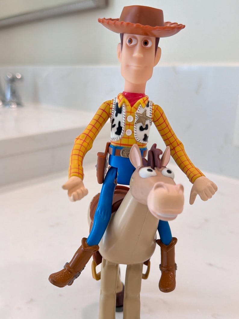 May include: A Woody action figure from the Toy Story franchise, wearing a brown cowboy hat, yellow plaid shirt, vest with cow print, blue trousers, and brown boots. The figure is riding a tan horse.