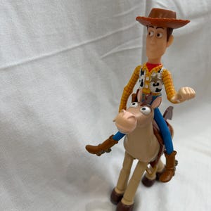 May include: A plastic toy figure of Woody from Toy Story riding a tan horse. Woody wears a brown cowboy hat, yellow shirt, vest, and blue trousers. The horse has brown boots and a blue saddle.