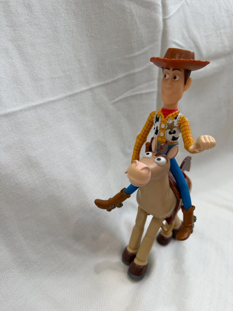 May include: A plastic toy figure of Woody from Toy Story riding a tan horse. Woody wears a brown cowboy hat, yellow shirt, vest, and blue trousers. The horse has brown boots and a blue saddle.