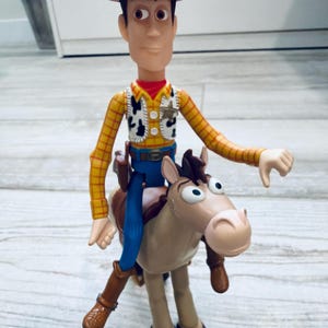 May include: A Woody action figure from the Toy Story franchise. The figure is wearing a brown cowboy hat, yellow plaid shirt, vest, blue trousers, and brown boots. Woody is riding a brown and tan horse.