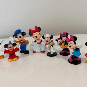 May include: A collection of Mickey and Minnie Mouse figurines. The figurines are in various poses and outfits, including classic Mickey in red shorts and Minnie in a polka-dot dress. The figurines are on a white surface.
