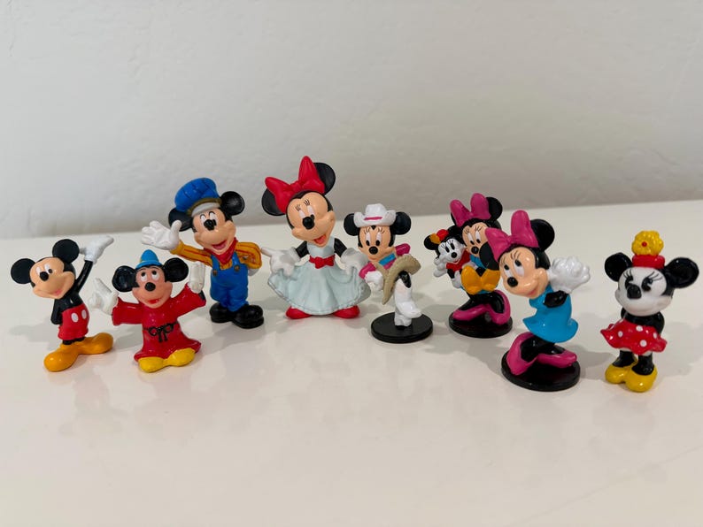 May include: A collection of Mickey and Minnie Mouse figurines. The figurines are in various poses and outfits, including classic Mickey in red shorts and Minnie in a polka-dot dress. The figurines are on a white surface.