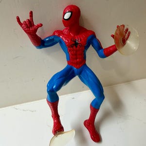 May include: A Spider-Man action figure in a dynamic pose, featuring a red and blue costume with a black spider emblem on the chest. The figure has a red mask with white eye lenses and is holding a clear disc. The figure is approximately 6 inches tall.