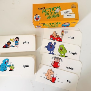 May include: A set of Easy Action Picture Words flash cards in an orange box, featuring illustrated cards with words like play, spin, stop, laugh, wade, and sip. Each card displays a colorful cartoon character performing an action next to the text.