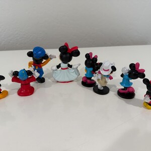 May include: A collection of small, colorful Mickey and Minnie Mouse figurines. The figurines feature various outfits and poses, with vibrant colors like red, blue, yellow, and black. The figures are arranged on a white surface.