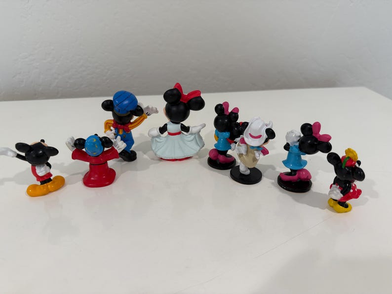 May include: A collection of small, colorful Mickey and Minnie Mouse figurines. The figurines feature various outfits and poses, with vibrant colors like red, blue, yellow, and black. The figures are arranged on a white surface.