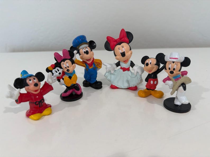 May include: A collection of seven small Mickey and Minnie Mouse figurines. The figurines feature Mickey and Minnie in various outfits, including a cowboy, a conductor, and a sorcerer. The figurines are brightly colored and made of plastic.