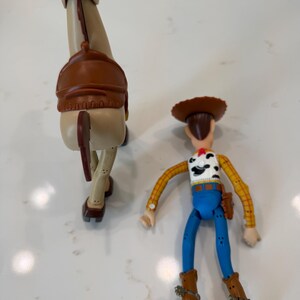 May include: Two plastic Toy Story figures: a tan horse with a brown saddle and a Woody action figure. Woody wears a cowboy hat, yellow plaid shirt, blue trousers, and brown boots. The horse has a brown tail.