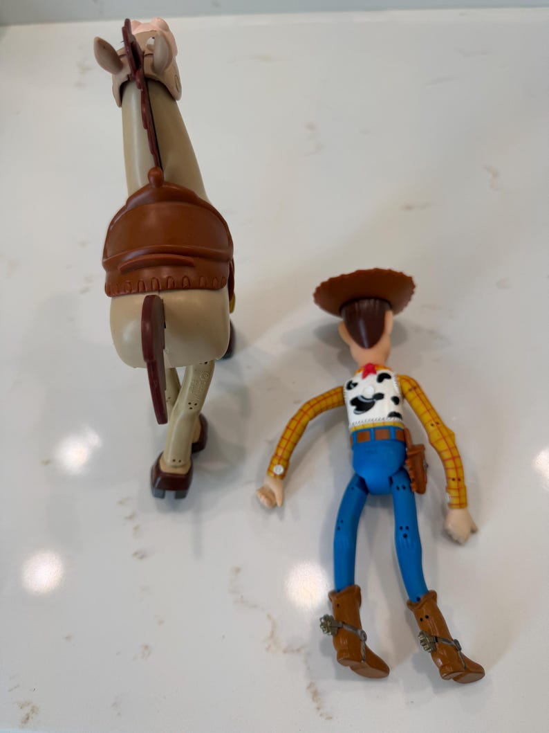 May include: Two plastic Toy Story figures: a tan horse with a brown saddle and a Woody action figure. Woody wears a cowboy hat, yellow plaid shirt, blue trousers, and brown boots. The horse has a brown tail.