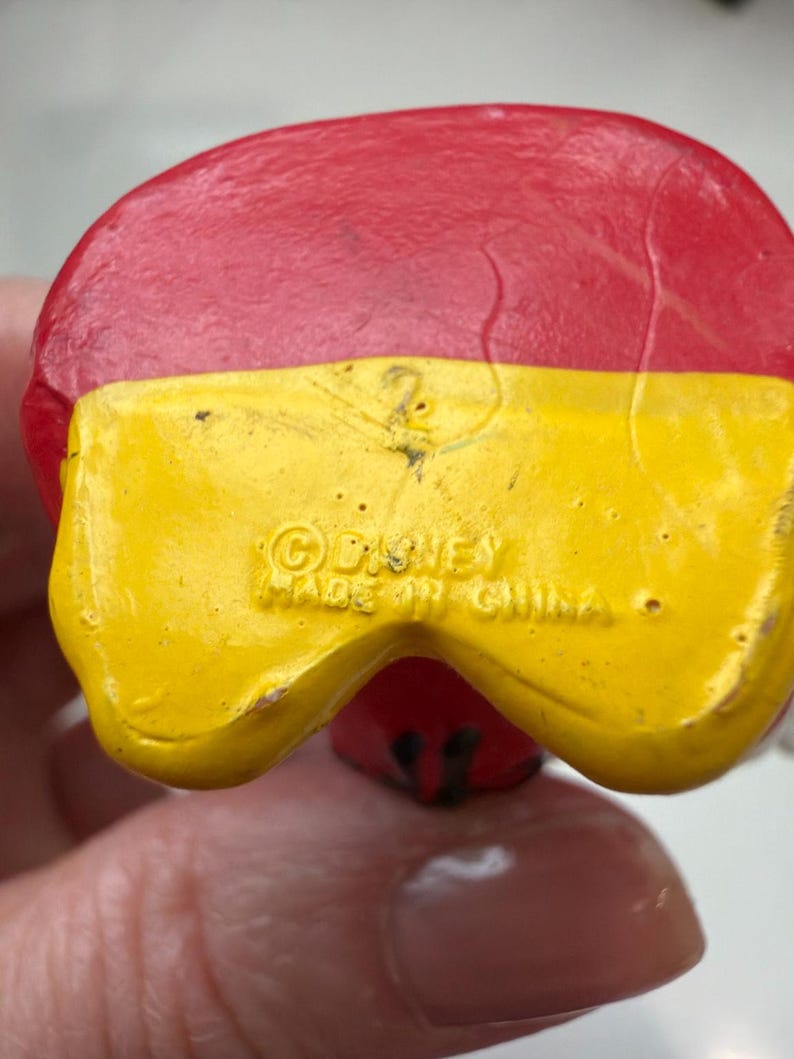May include: Close-up of a red and yellow figurine. The figurine's base is yellow with the text "&copy; Disney Made in China". The top is red. The figurine is a collectible item.