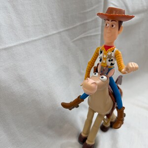 May include: A plastic toy figure of Woody from Toy Story, wearing a brown cowboy hat, yellow shirt, and blue trousers. He is riding a tan horse with brown boots. The toy is set against a white background.