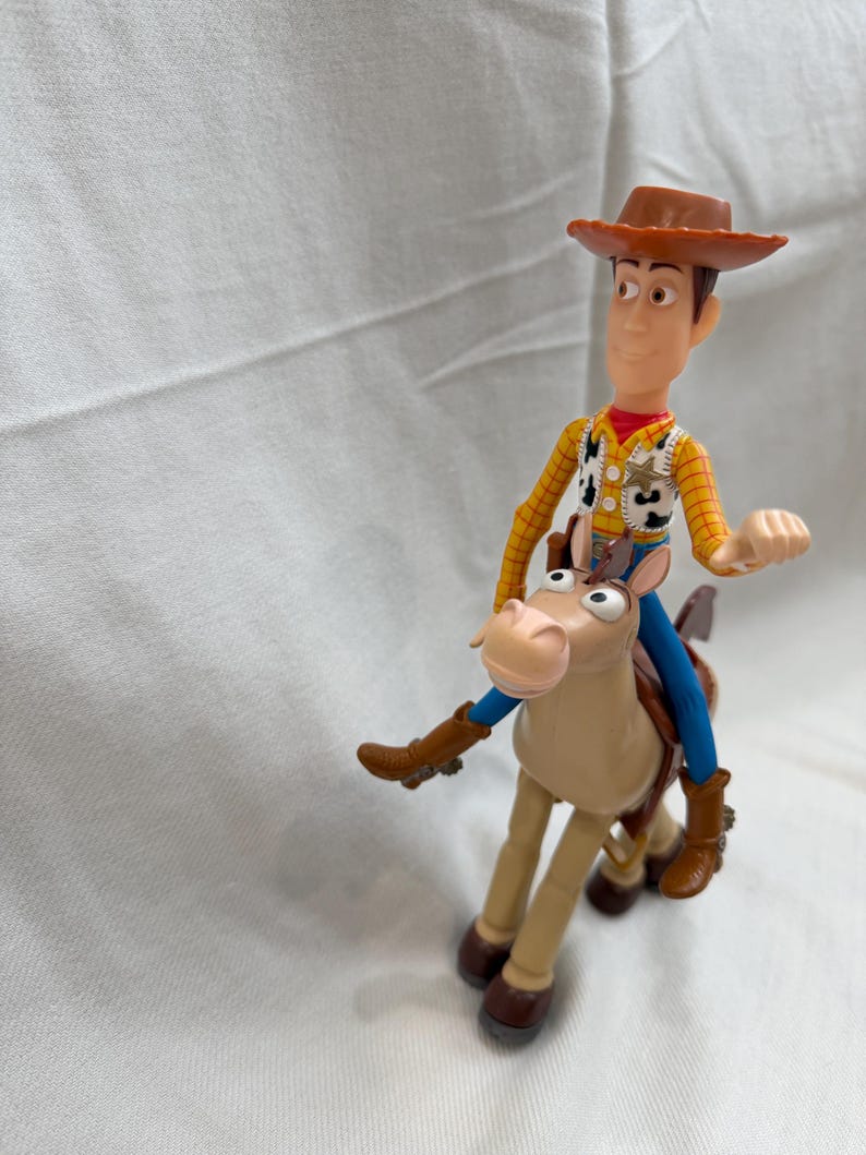 May include: A plastic toy figure of Woody from Toy Story, wearing a brown cowboy hat, yellow shirt, and blue trousers. He is riding a tan horse with brown boots. The toy is set against a white background.