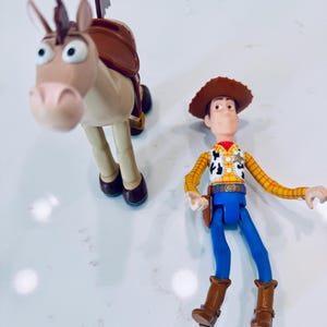 May include: Two plastic Toy Story figures: Woody, a cowboy with a brown hat, yellow shirt, blue trousers, and brown boots, and Bullseye, a tan horse with a brown saddle. The figures are on a white surface.