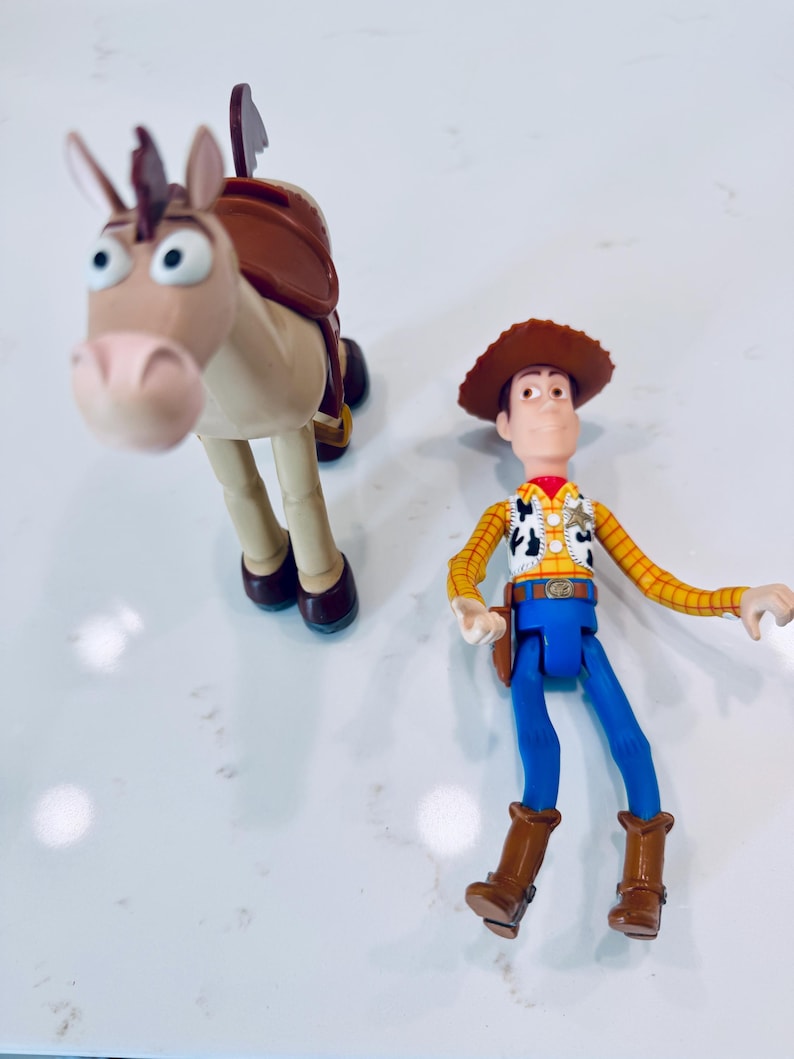May include: Two plastic Toy Story figures: Woody, a cowboy with a brown hat, yellow shirt, blue trousers, and brown boots, and Bullseye, a tan horse with a brown saddle. The figures are on a white surface.