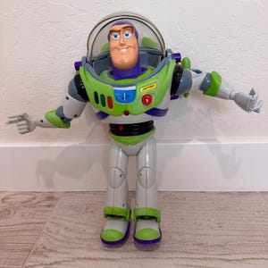 May include: A Buzz Lightyear action figure from the Toy Story franchise. The figure is primarily white, green, and purple, with a clear plastic helmet. The figure has its arms outstretched and is standing on a wooden floor.