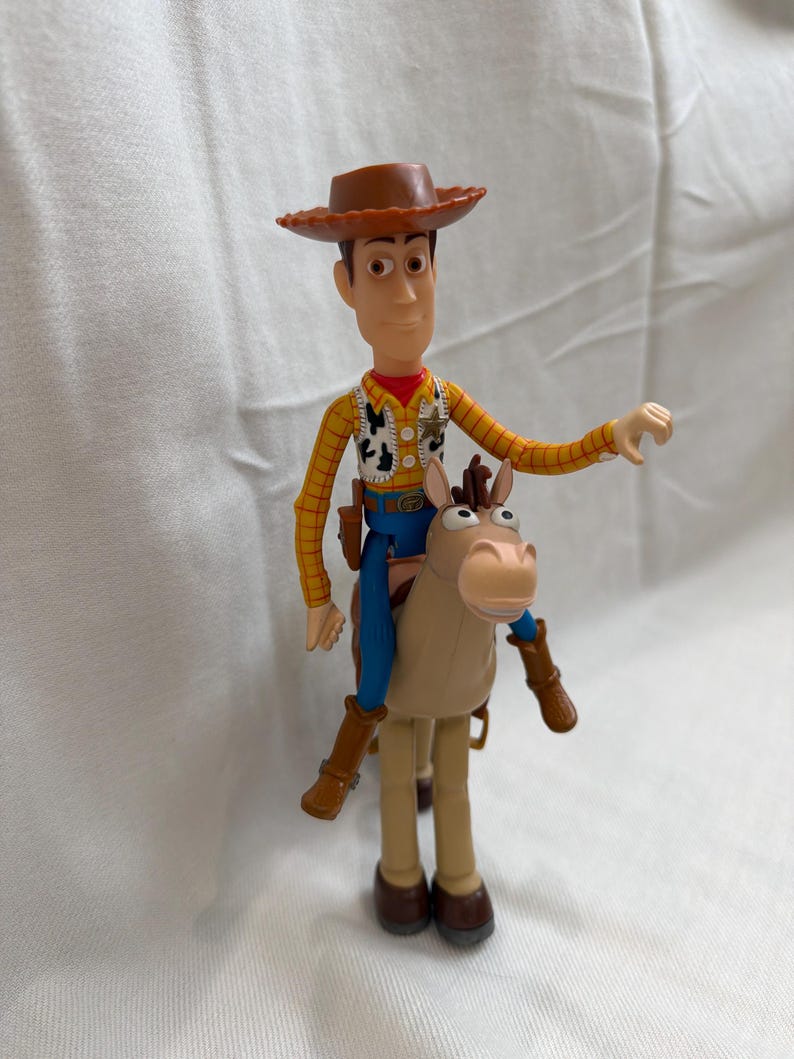 May include: A Woody action figure riding a tan horse. Woody wears a brown cowboy hat, a yellow plaid shirt, a vest, blue trousers, and brown boots. The horse has a tan body, brown legs, and a light brown face.