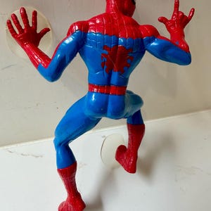 May include: A plastic Spider-Man action figure in a climbing pose, viewed from the back. The figure is predominantly blue and red, with a spider emblem on the back. The figure has suction cups attached to the hands and feet.