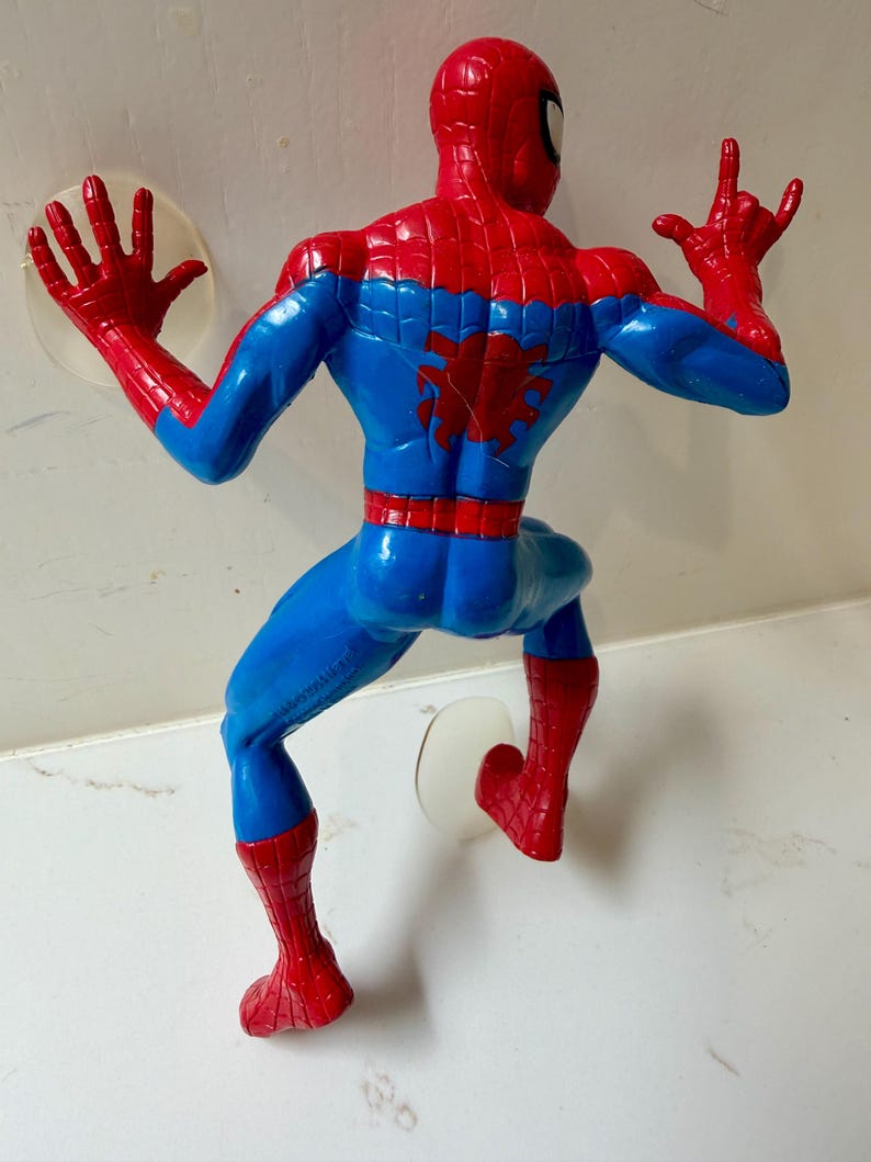 May include: A plastic Spider-Man action figure in a climbing pose, viewed from the back. The figure is predominantly blue and red, with a spider emblem on the back. The figure has suction cups attached to the hands and feet.