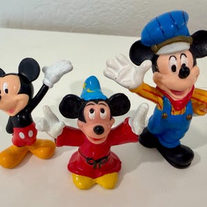 May include: Three Mickey Mouse figurines in various poses. One figure is dressed in a blue conductor's outfit, another in a red sorcerer's robe, and the third in a classic black and red outfit. All have white gloves and yellow shoes.