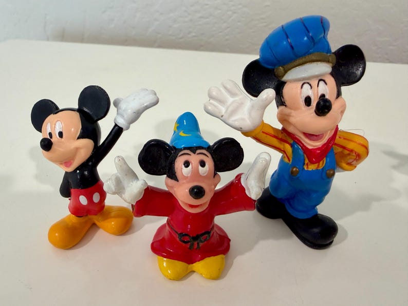 May include: Three Mickey Mouse figurines in various poses. One figure is dressed in a blue conductor's outfit, another in a red sorcerer's robe, and the third in a classic black and red outfit. All have white gloves and yellow shoes.