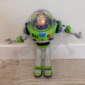 May include: A Buzz Lightyear action figure, featuring a green and white spacesuit with purple accents. The figure has a clear helmet, a smiling face, and outstretched arms. The word "LIGHTYEAR" is visible on the chest.