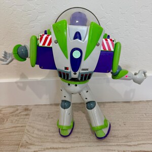May include: A Buzz Lightyear action figure, featuring a white and green spacesuit with purple accents. The toy has a clear helmet, articulated arms, and is posed with arms outstretched. The figure is standing on a light-colored wooden floor.