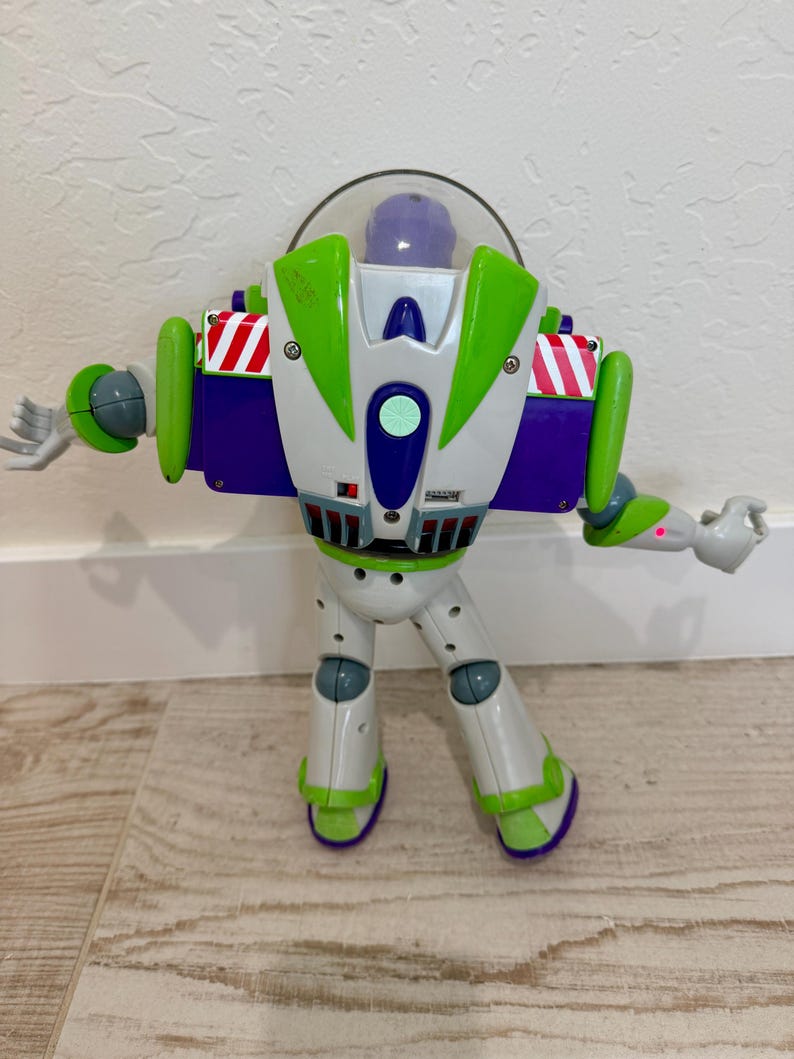 May include: A Buzz Lightyear action figure, featuring a white and green spacesuit with purple accents. The toy has a clear helmet, articulated arms, and is posed with arms outstretched. The figure is standing on a light-colored wooden floor.