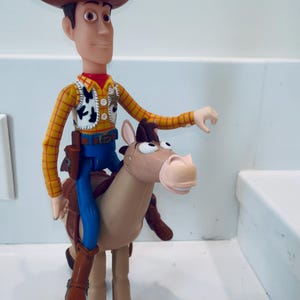 May include: A Woody action figure from the Toy Story franchise. The figure is wearing a brown cowboy hat, yellow shirt, vest, blue trousers, and brown boots. Woody is riding a tan horse.