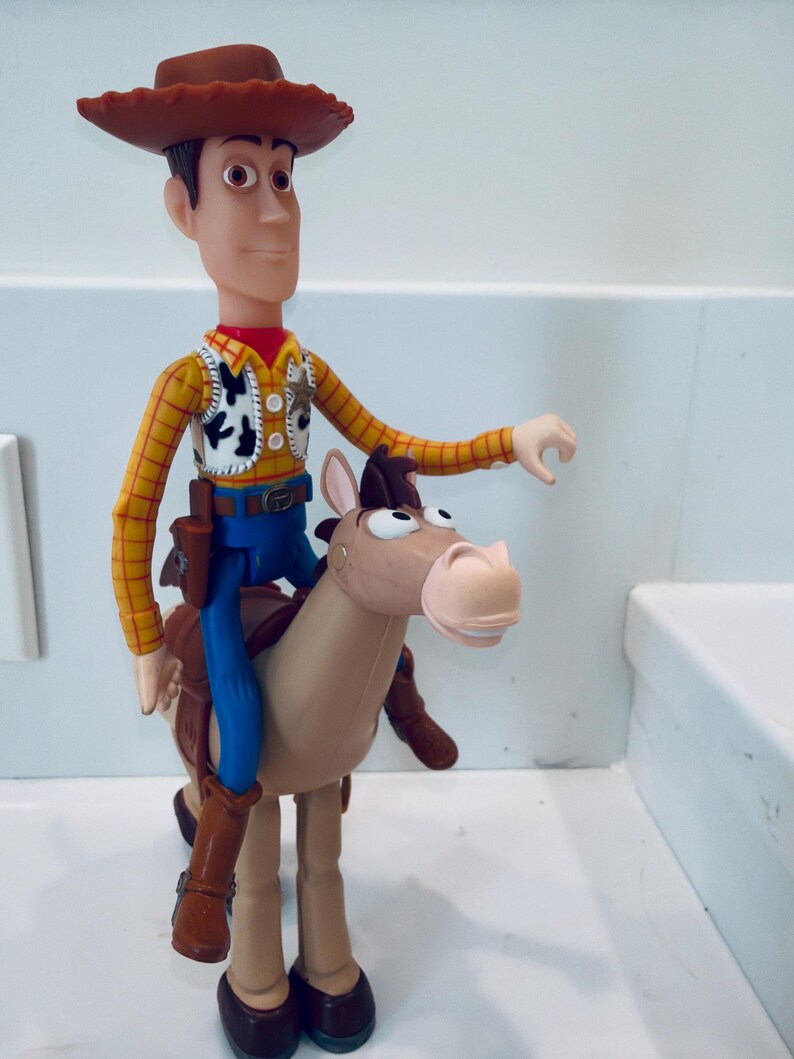May include: A Woody action figure from the Toy Story franchise. The figure is wearing a brown cowboy hat, yellow shirt, vest, blue trousers, and brown boots. Woody is riding a tan horse.