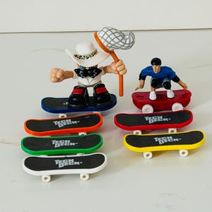 May include: A collection of Tech Deck fingerboards and two small plastic figures. One figure wears a white hat and holds a net, while the other wears a black helmet and blue shirt. The skateboards feature black grip tape with the Tech Deck logo.