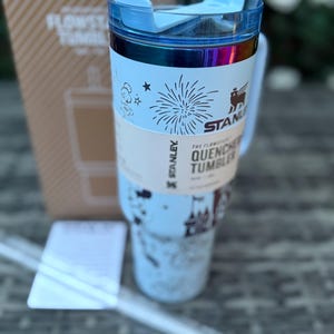 May include: A light blue Stanley Flowstate Quencher tumbler with a clear straw. The tumbler has a blue lid and a white band with the text "The Flowstate Quencher Tumbler" and a fireworks graphic. The Stanley logo is visible on the tumbler and the box.