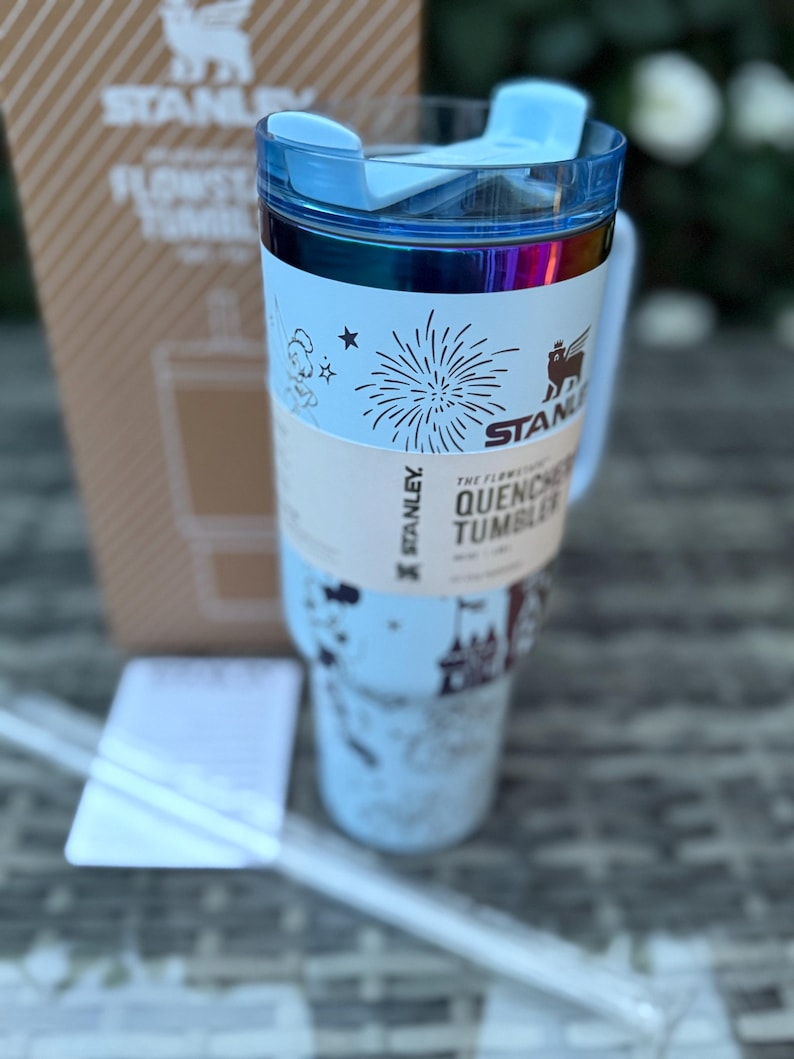 May include: A light blue Stanley Flowstate Quencher tumbler with a clear straw. The tumbler has a blue lid and a white band with the text "The Flowstate Quencher Tumbler" and a fireworks graphic. The Stanley logo is visible on the tumbler and the box.