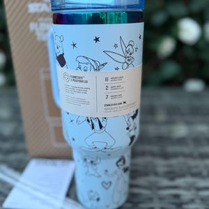 May include: A light blue insulated tumbler with a blue lid and handle. The tumbler features black and white illustrations of Disney characters. A paper band around the tumbler has text that reads "FLOWSTATE 3-POSITION LID" and "HOURS COLD".