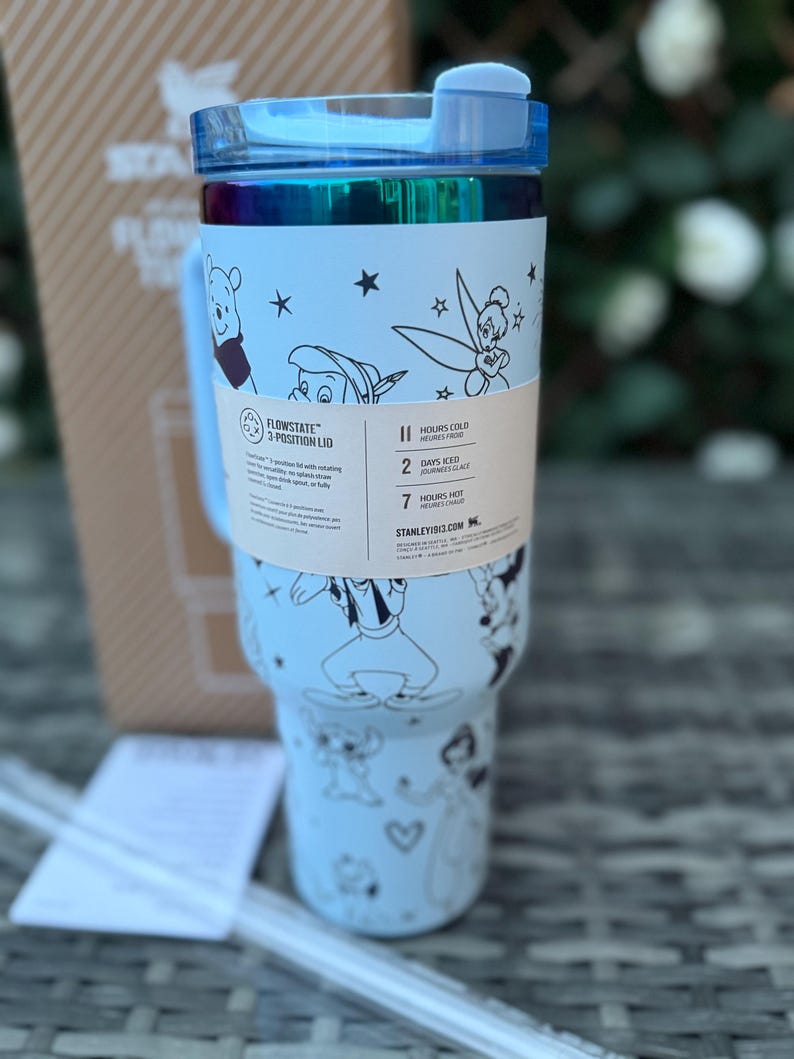 May include: A light blue insulated tumbler with a blue lid and handle. The tumbler features black and white illustrations of Disney characters. A paper band around the tumbler has text that reads "FLOWSTATE 3-POSITION LID" and "HOURS COLD".