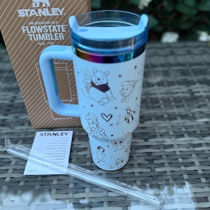May include: A white Stanley Flowstate tumbler with a light blue handle and lid. The tumbler features a wraparound design with cartoon characters. A clear straw and the product box are also visible.