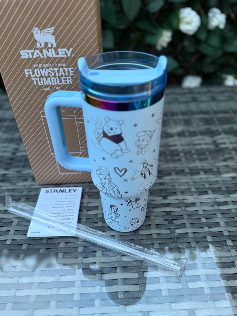 May include: A white Stanley Flowstate tumbler with a light blue handle and lid. The tumbler features a wraparound design with cartoon characters. A clear straw and the product box are also visible.