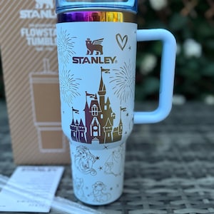 May include: A white Stanley Flowstate tumbler with a light blue handle and lid. The tumbler features a castle design, fireworks, and character illustrations. The Stanley logo is visible. Includes two clear straws.