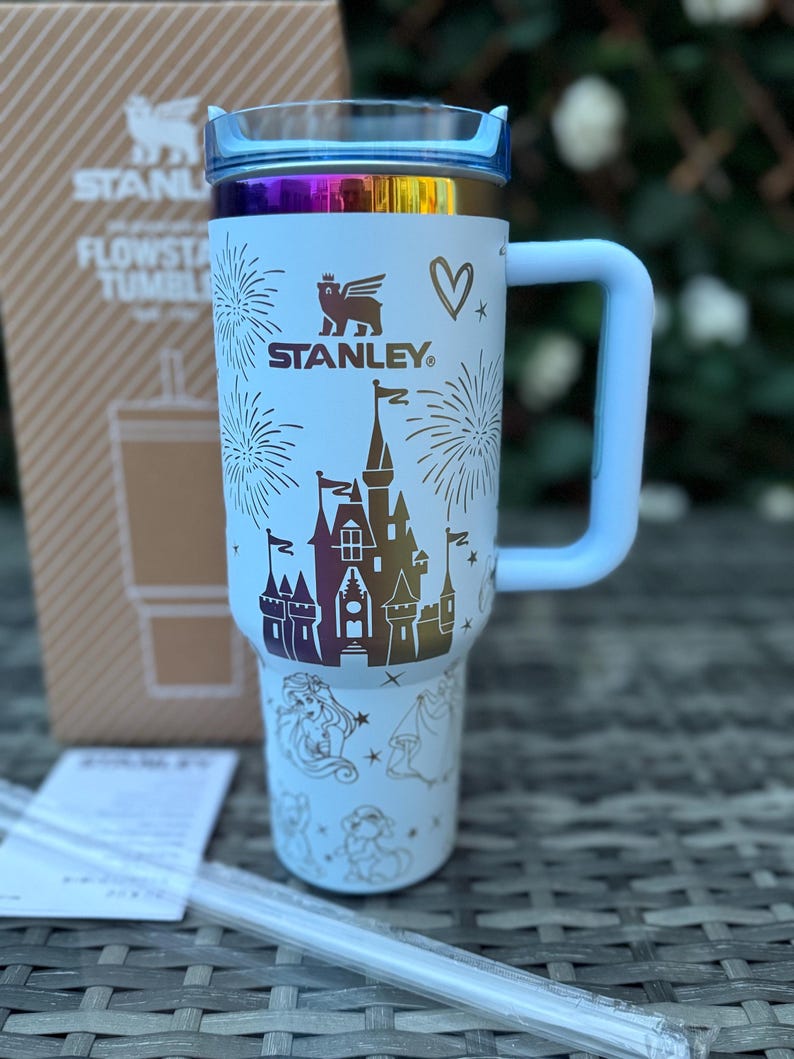 May include: A white Stanley Flowstate tumbler with a light blue handle and lid. The tumbler features a castle design, fireworks, and character illustrations. The Stanley logo is visible. Includes two clear straws.