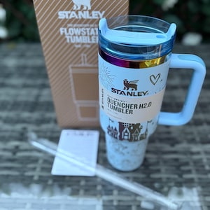 May include: A light blue Stanley Quencher H2.0 Flowstate Tumbler with a handle, lid, and straw. The tumbler has a white label with the text "QUENCHER H2.0 TUMBLER" and a decorative design. A cardboard box with the Stanley logo is in the background.