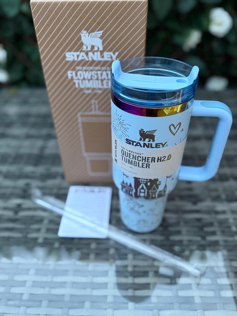 May include: A light blue Stanley Quencher H2.0 Flowstate Tumbler with a handle, lid, and straw. The tumbler has a white label with the text "QUENCHER H2.0 TUMBLER" and a decorative design. A cardboard box with the Stanley logo is in the background.