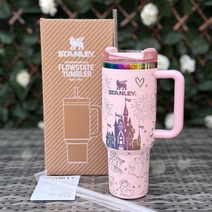 Peut inclure: Un gobelet Stanley Flowstate rose avec une poignée, une paille et un couvercle. Le gobelet présente un château et des personnages. La boîte à côté porte le logo Stanley et des informations sur le produit. Une paille transparente et un petit papier sont également visibles.