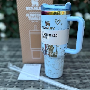 May include: A light blue Stanley Quencher H2.0 tumbler with a handle, straw, and a cardboard box. The tumbler features a light blue lid, a gold rim, and a light blue body with a castle and heart design. The box has the Stanley logo.