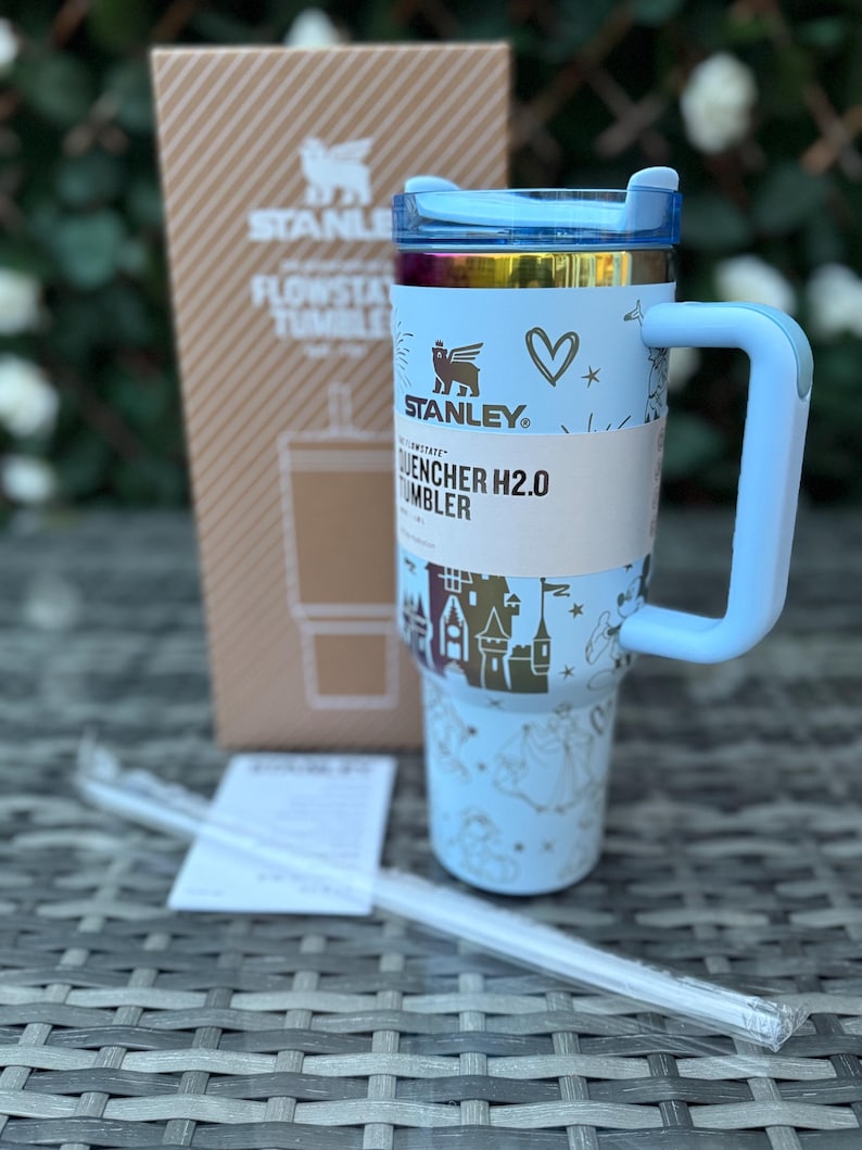 May include: A light blue Stanley Quencher H2.0 tumbler with a handle, straw, and a cardboard box. The tumbler features a light blue lid, a gold rim, and a light blue body with a castle and heart design. The box has the Stanley logo.