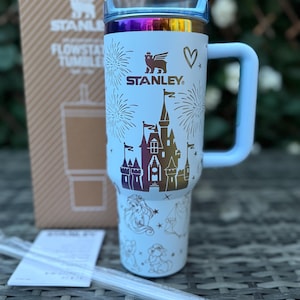 May include: A light blue Stanley Flowstate tumbler with a handle, featuring a castle design and the Stanley logo. The lid has a rainbow iridescent finish. The tumbler is accompanied by a box and a straw.