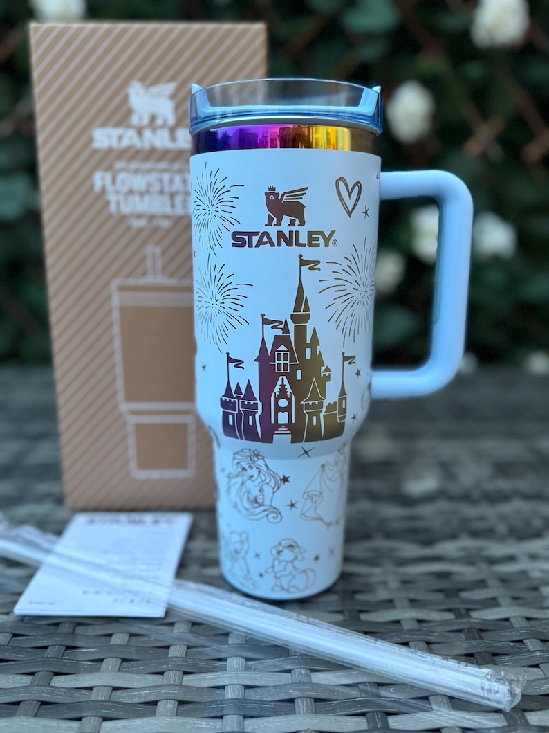 May include: A light blue Stanley Flowstate tumbler with a handle, featuring a castle design and the Stanley logo. The lid has a rainbow iridescent finish. The tumbler is accompanied by a box and a straw.