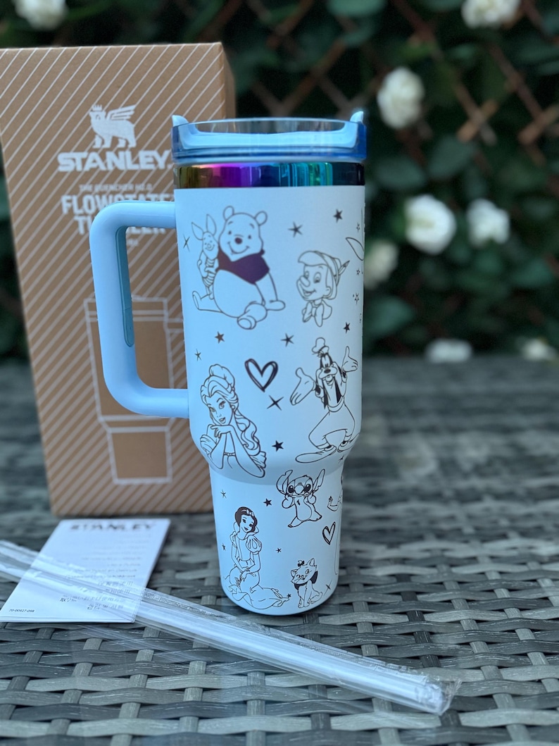 May include: A light blue Stanley Flowstate tumbler with a handle and a rainbow-colored lid. The tumbler features black line drawings of Disney characters, including Winnie the Pooh, and a clear straw.